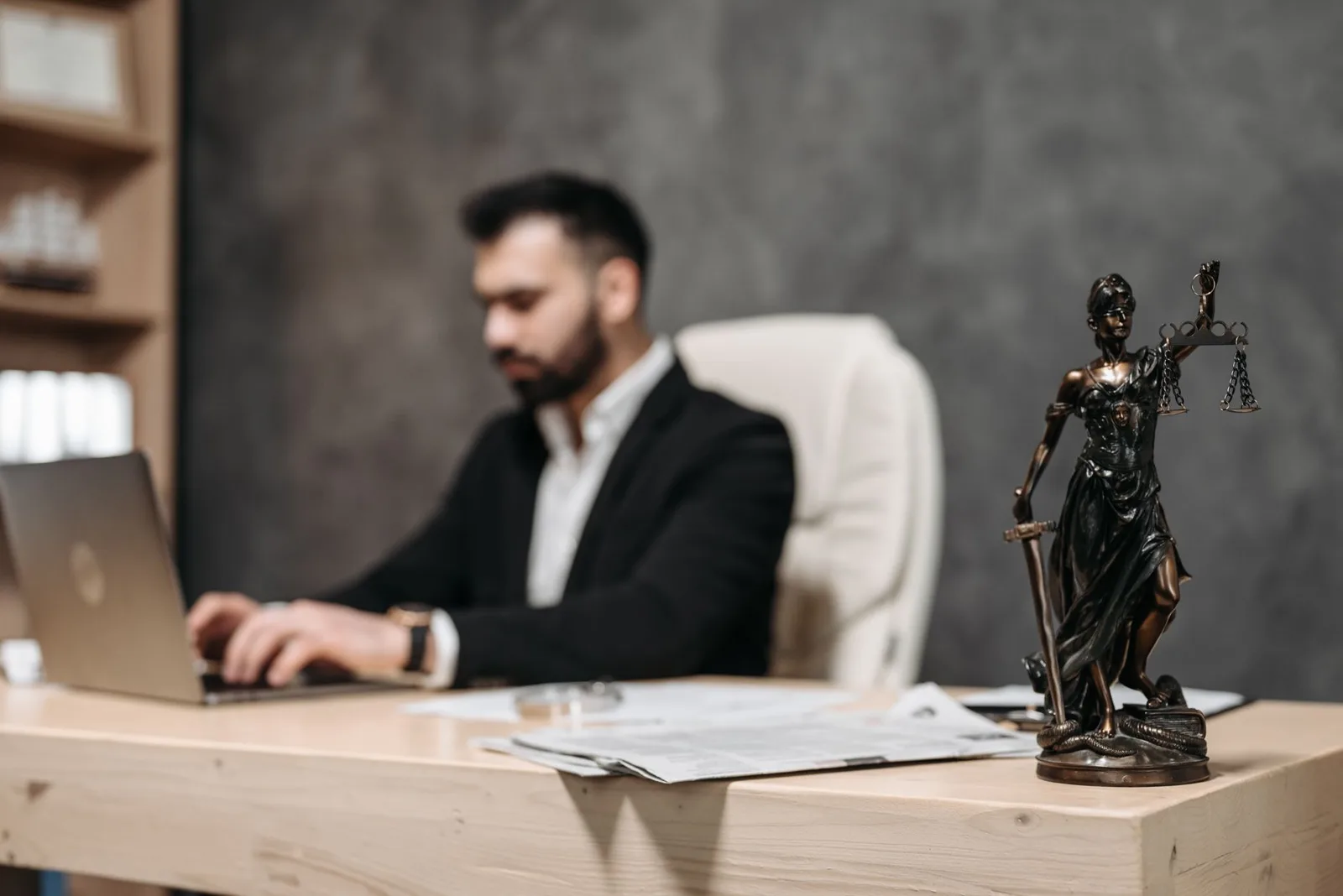 A businessman working on a laptop in an office with a Lady Justice statue in focus on the desk.