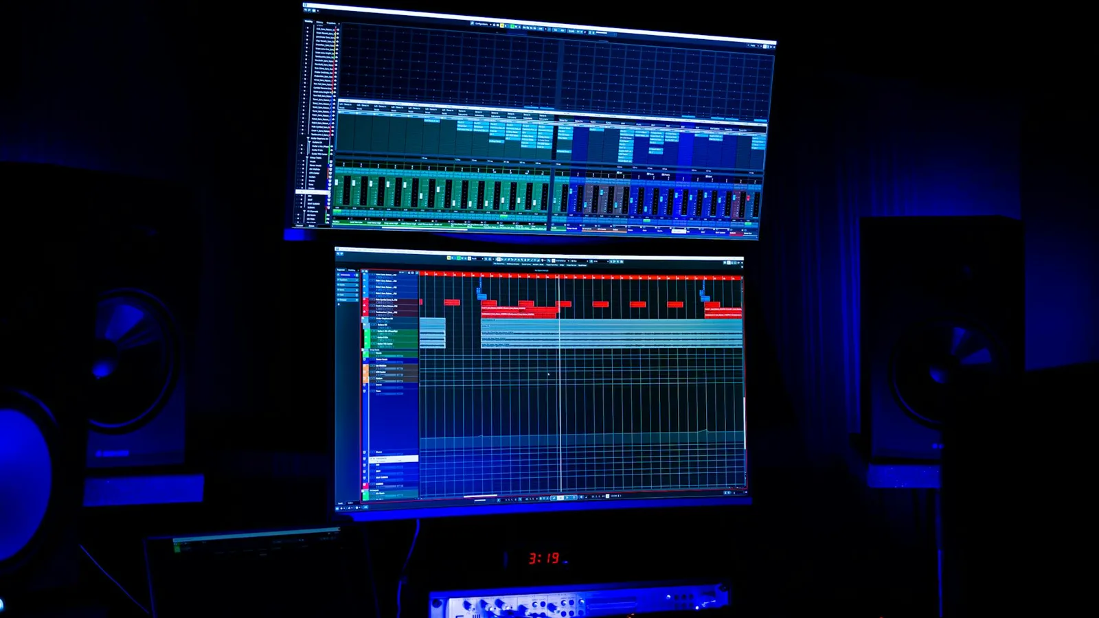 A modern recording studio featuring two screens displaying audio editing software under blue neon lighting.