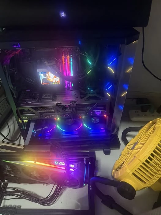 KoishiAI's actual benchmark rig — open-case dual-GPU workstation with RTX 5090 (top) and Zotac RTX 5080 (bottom), RGB-lit, with a supplementary desk fan for airflow. Photographed in Thailand.